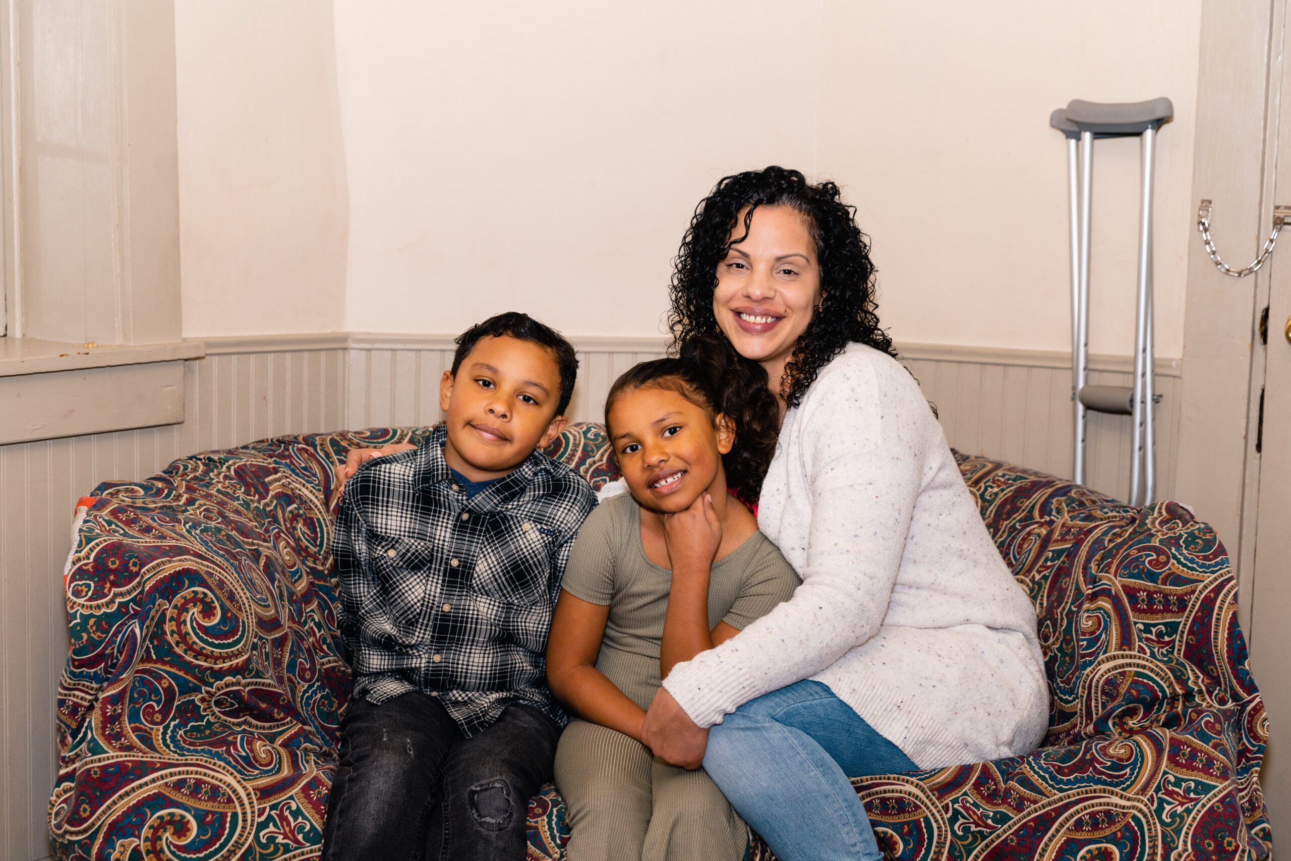 Woman sitting on a patterned couch with her two children. A pair of crutches lean against the wall.