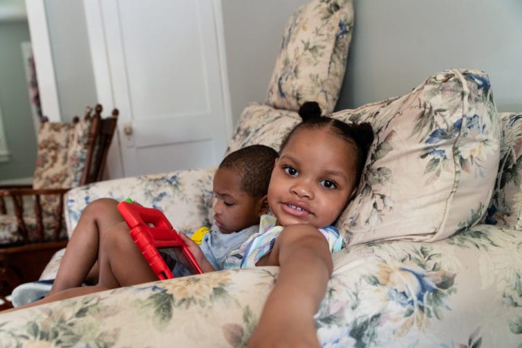 Two children sit on a loveseat with an electronic device