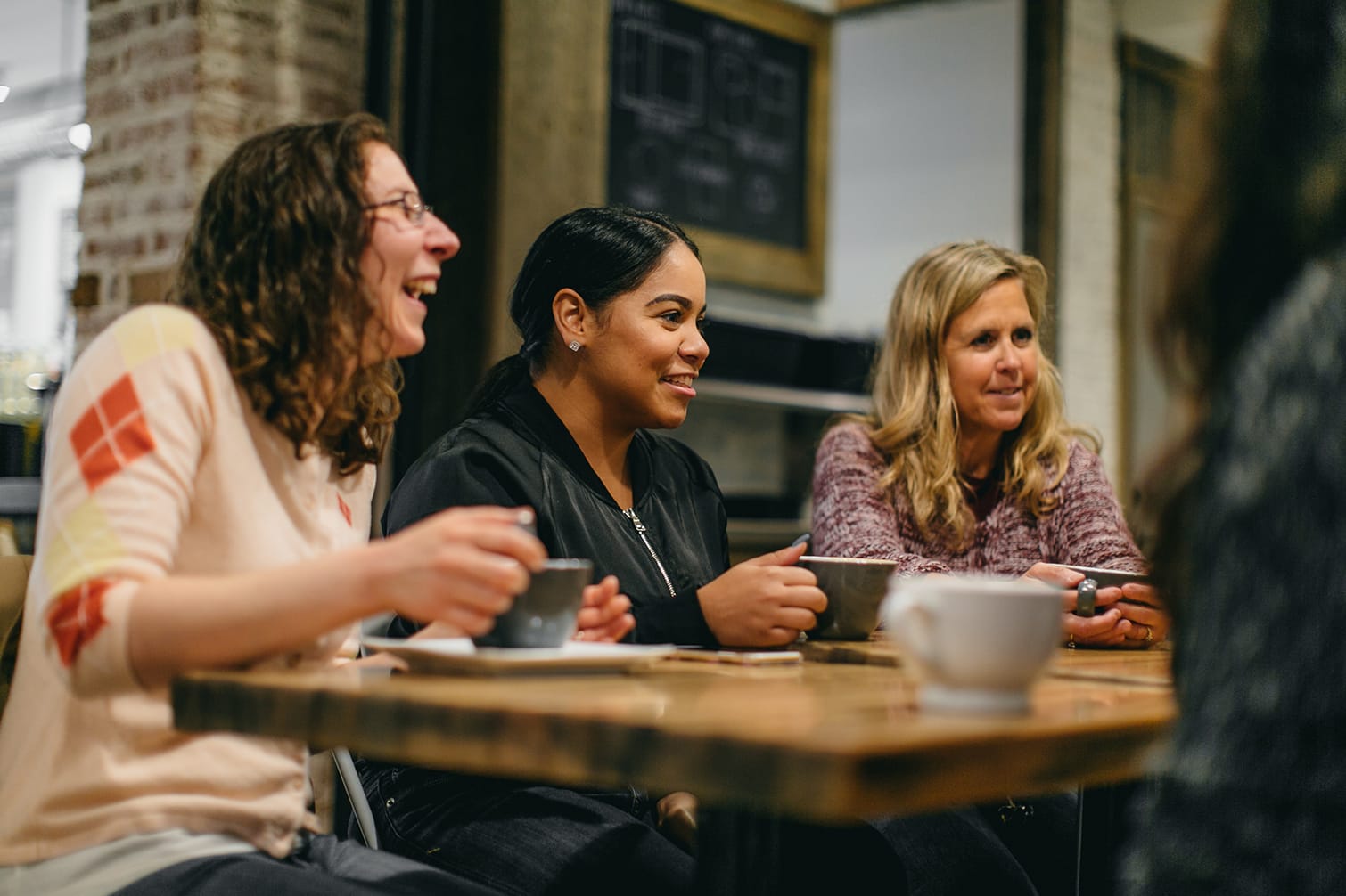 three happy women sit beside each other at a coffee shop with coffee mugs